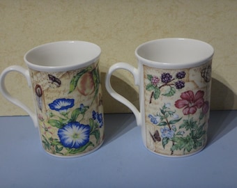 May include: Two white ceramic mugs with floral designs. The mug on the left has a peach, butterflies, and blue flowers. The mug on the right has blackberries, a pink flower, and a butterfly.