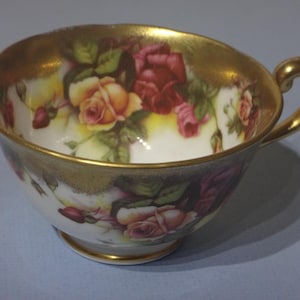 Op de afbeelding: Een wit theekopje met een gouden rand en een bloemenmotief. Het kopje heeft een delicaat patroon van roze, gele en rode rozen met groene bladeren. Het kopje heeft een handvat en een kleine voet.