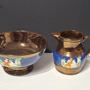 May include: A vintage ceramic bowl and creamer set. The bowl has a bronze interior and a blue exterior with a decorative scene. The creamer is bronze with a blue band and a matching scene. A classic tableware set.