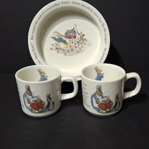 Puede incluir: Un plato y dos tazas de cerámica color crema con ilustraciones de conejos vestidos. El plato tiene un borde con texto y las tazas tienen asas. Las ilustraciones son en colores suaves.