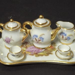 Peut inclure: Service à thé miniature en porcelaine avec bordure dorée. L'ensemble comprend une théière, un pot à crème, un sucrier, deux tasses et soucoupes, le tout sur un plateau décoratif. Des motifs floraux violets et bleus ornent la porcelaine blanche.
