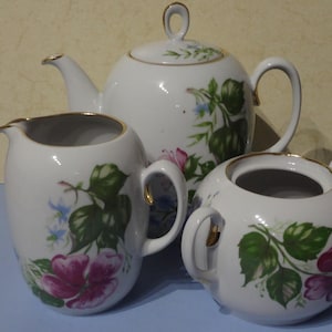 Könnte beinhalten: Ein weißes Porzellan-Tee-Set mit floralem Design. Das Set beinhaltet eine Teekanne, eine Milchkännchen und eine Zuckerdose. Die Teekanne hat einen goldenen Griff und Ausguss. Die Milchkännchen und die Zuckerdose haben goldene Ränder.