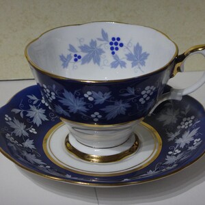 Puede incluir: Un juego de taza y platillo de té azul y blanco con un diseño de vid y uvas. La taza y el platillo tienen un borde dorado.