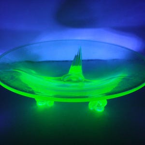 May include: A bright green glass dish with a central point and three feet. The dish is made of uranium glass and glows under a black light.