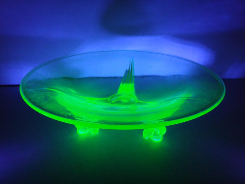May include: A bright green glass dish with a central point and three feet. The dish is made of uranium glass and glows under a black light.