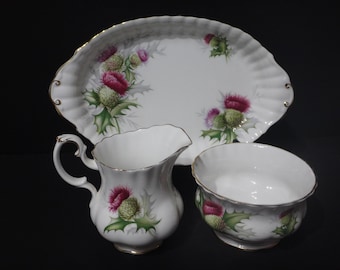 May include: A white porcelain serving set featuring a platter, creamer, and sugar bowl. Each piece is decorated with a thistle motif in shades of pink, green, and gold. The set has a scalloped edge and gold trim.