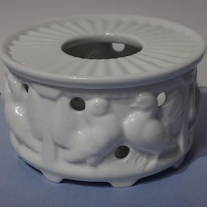May include: White ceramic tea warmer with a decorative design of birds and leaves. The warmer has a round top with a hole in the centre for a teapot.