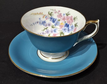 May include: A teal teacup and saucer set. The teacup features a floral design in pink, blue, and purple on a white interior. The rim and handle are trimmed with gold. The saucer is teal with a gold rim.