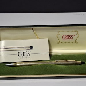 Könnte beinhalten: Ein goldfarbener Cross-Stift in einer schwarzen Präsentationsbox mit grünem Samtinnenraum. Die Box enthält auch eine weiße Karte mit einer silbernen Stiftillustration und dem Cross-Logo. Ein klassisches Schreibgerät.
