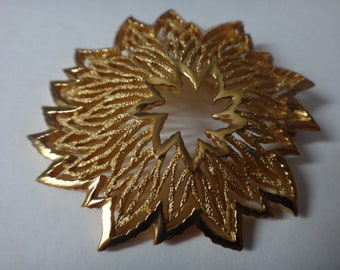 May include: A gold-toned metal brooch in the shape of a stylized flower with a circular opening in the center.