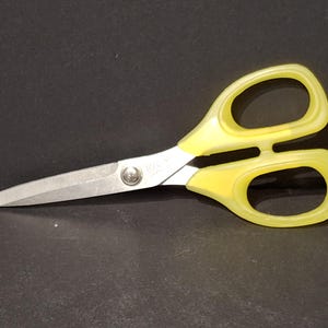 Może przedstawiać: Para nożyczek z żółtą rączką i srebrnymi ostrzami. Nożyczki leżą na ciemnej powierzchni. Uchwyty mają zaokrągloną konstrukcję ułatwiającą chwyt.