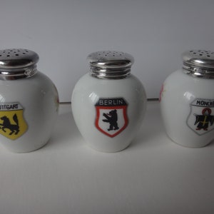 May include: Three white ceramic salt shakers with silver tops. Each shaker features a different city crest: Stuttgart, Berlin, and Munich.