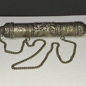Könnte beinhalten: Ein silberfarbener, zylindrischer Behälter mit aufwendigen floralen und geometrischen Mustern. Der Behälter hat spitze Enden und ist an zwei Ketten aufgehängt. Das Design deutet darauf hin, dass es sich um einen dekorativen oder zeremoniellen Gegenstand handeln könnte.
