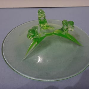 May include: A green glass pedestal dish with three leaf-shaped feet. The dish is made of uranium glass and glows under a black light.