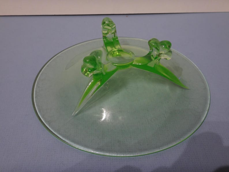 May include: A green glass pedestal dish with three leaf-shaped feet. The dish is made of uranium glass and glows under a black light.
