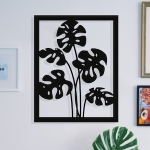 Puede incluir: Escena de salón con un cuadro negro con diseño de hojas de monstera. Un sofá de terciopelo azul con cojines es visible. Una máquina de escribir vintage está sobre una mesa de madera. Otras obras de arte en la pared.