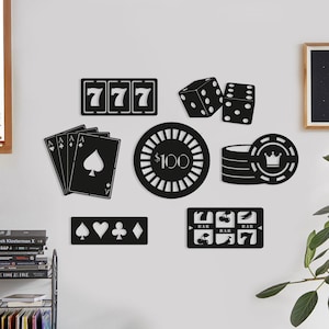 Puede incluir: Arte de pared de metal negro con diseños temáticos de casino. Incluye una máquina tragamonedas, naipes, dados, fichas de póquer y una ficha de $100. El arte se exhibe en una pared blanca, creando un contraste audaz.