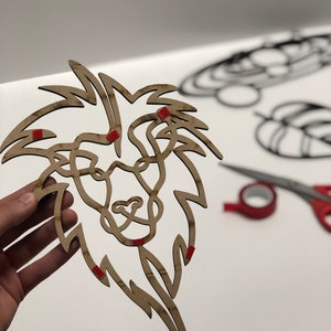 以下が含まれることがあります： 詳細な鬣と顔を持つ、木製のライオンの頭のウォールアート。ライオンは薄茶色で、赤い粘着性の正方形が付いています。他の装飾的な形とハサミが背景にあります。
