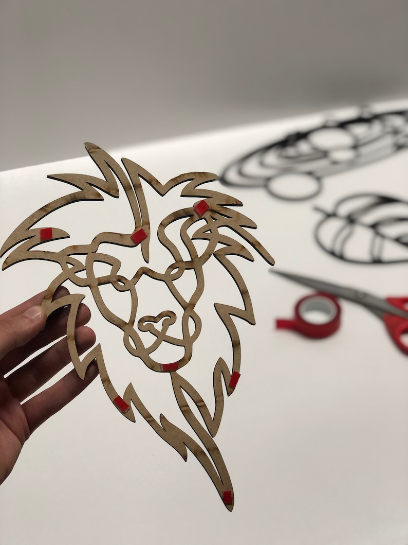 以下が含まれることがあります： 詳細な鬣と顔を持つ、木製のライオンの頭のウォールアート。ライオンは薄茶色で、赤い粘着性の正方形が付いています。他の装飾的な形とハサミが背景にあります。