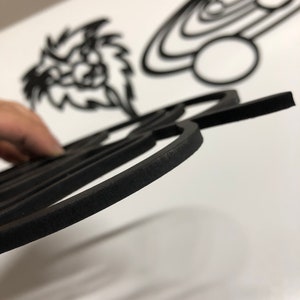 以下が含まれることがあります： 黒いレーザーカットの木製アート作品のクローズアップ。前景には、丸みを帯びたエッジを持つ詳細な幾何学模様があります。背景には、様式化されたライオンの頭や惑星のデザインなど、他の切り抜かれた形が見えます。作品は白い表面にあります。