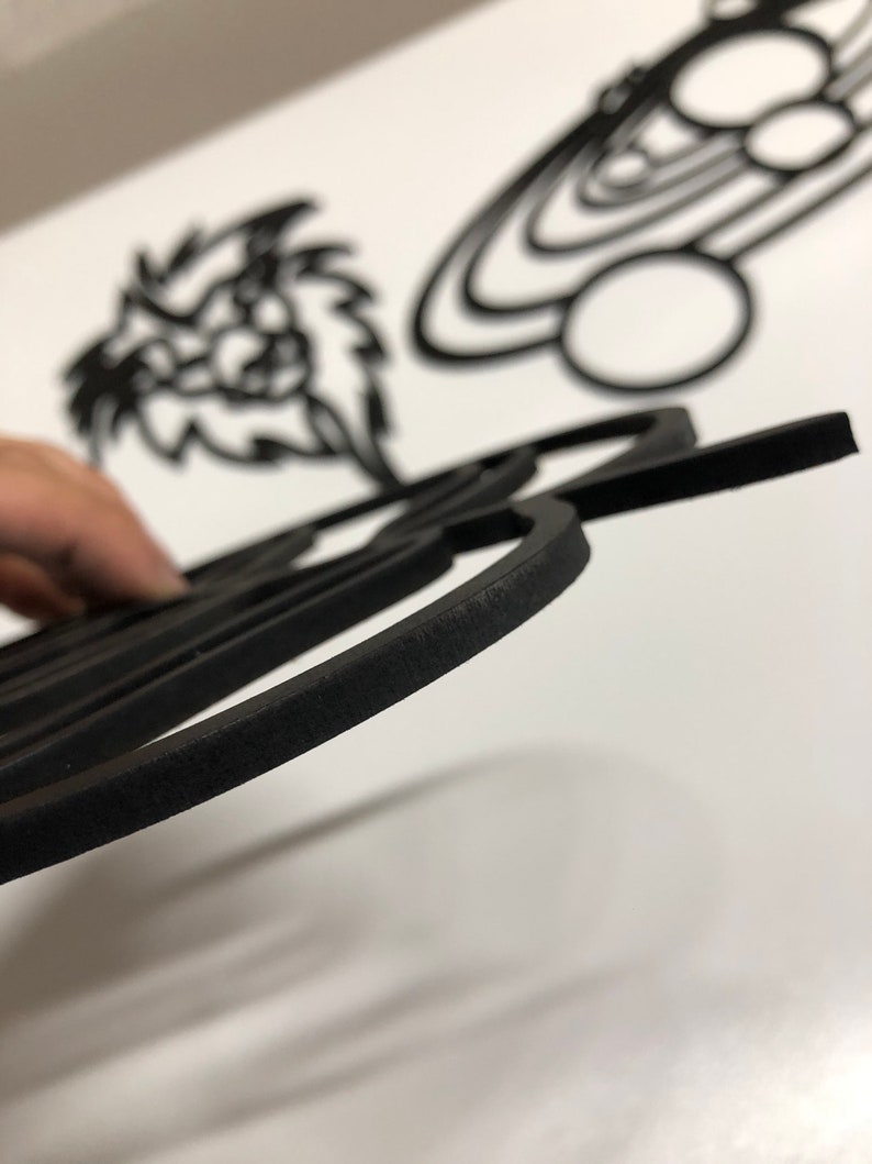 以下が含まれることがあります： 黒いレーザーカットの木製アート作品のクローズアップ。前景には、丸みを帯びたエッジを持つ詳細な幾何学模様があります。背景には、様式化されたライオンの頭や惑星のデザインなど、他の切り抜かれた形が見えます。作品は白い表面にあります。