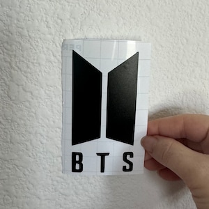 Puede incluir: Pegatina de vinilo en blanco y negro con el logotipo de BTS y las letras "BTS" debajo.