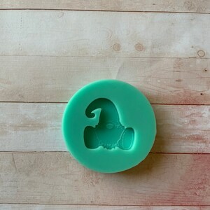 May include: A green silicone mold for making a gnome figurine. The mold is round and has a raised design of a gnome with a pointed hat, beard, and arms.