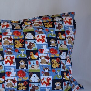 May include: A rectangular pillowcase featuring a vibrant pattern of cartoon dogs, ducks, and other animals arranged in a grid. The background consists of blue, black, and white squares. The pillowcase rests on a white surface.