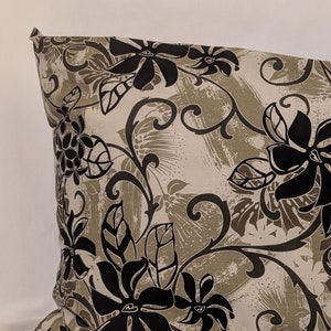 May include: Rectangular pillow with a floral pattern. The pillow features large black flowers and leaves against a beige and gray background. The pillow is placed on a white surface.