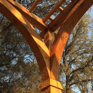 Puede incluir: Una pérgola de madera con un techo de celosía. La pérgola está hecha de madera natural de color marrón cálido. La pérgola está sostenida por gruesas vigas de madera que están curvadas en la parte superior. Las vigas se unen en la parte superior para formar una forma triangular. La pérgola está rodeada de árboles.