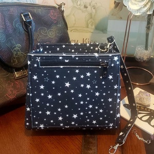Puede incluir: Un bolso bandolera negro con un patrón de estrellas blancas. El bolso tiene un bolsillo con cremallera y una correa larga ajustable.