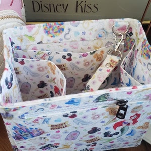 May include: White fabric organizer with a zipper closure and multiple interior compartments. The organizer is decorated with a colorful pattern of Disney characters, including Mickey Mouse, Cinderella's glass slipper, and castles. A keychain with the text "Mickey Forever" is attached.