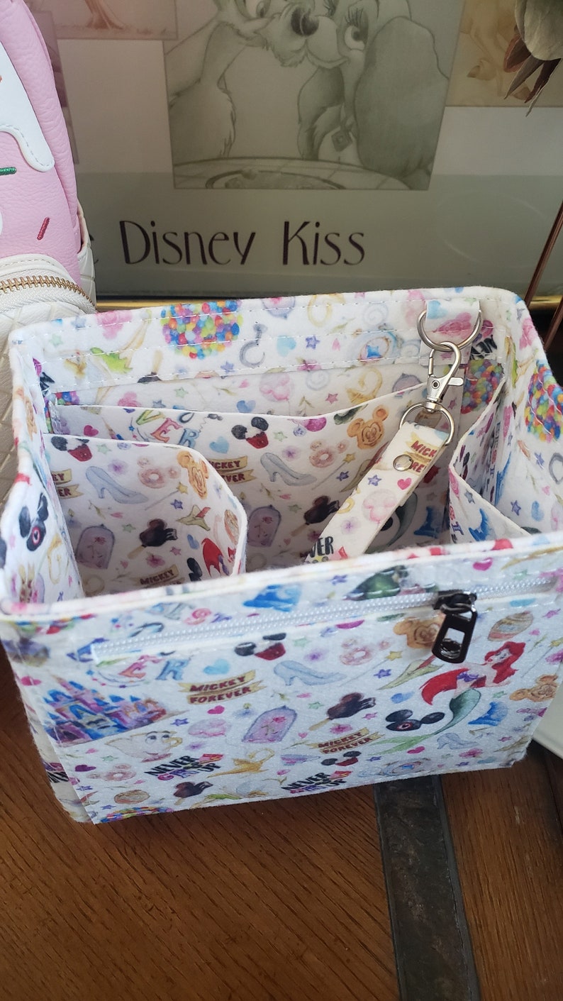 May include: White fabric organizer with a zipper closure and multiple interior compartments. The organizer is decorated with a colorful pattern of Disney characters, including Mickey Mouse, Cinderella's glass slipper, and castles. A keychain with the text "Mickey Forever" is attached.