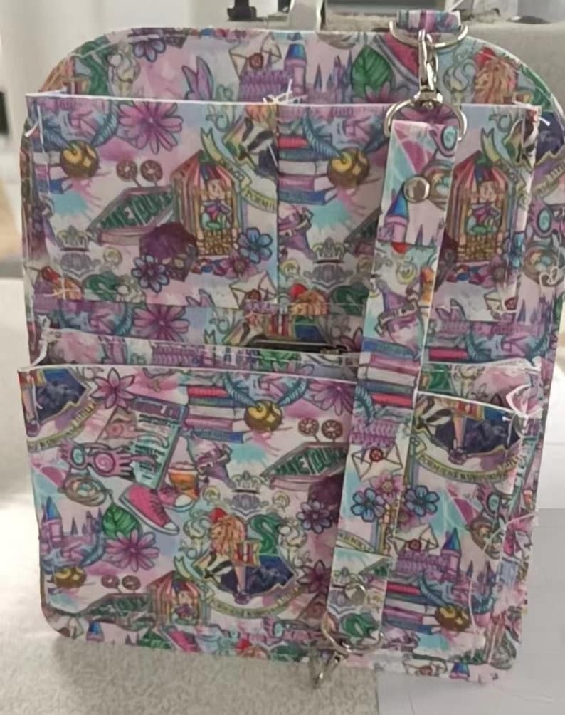 May include: A multi-pocket backpack with a colorful pattern featuring castles, books, and floral designs. The bag has a detachable strap and silver-tone hardware. The color palette includes pink, purple, and green.