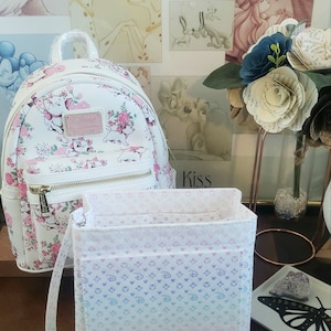 May include: White backpack with a Marie the Cat design, featuring pink flowers and a gold zipper. A matching organizer with a gradient pattern and a detachable strap sits in front. The background includes Disney art.