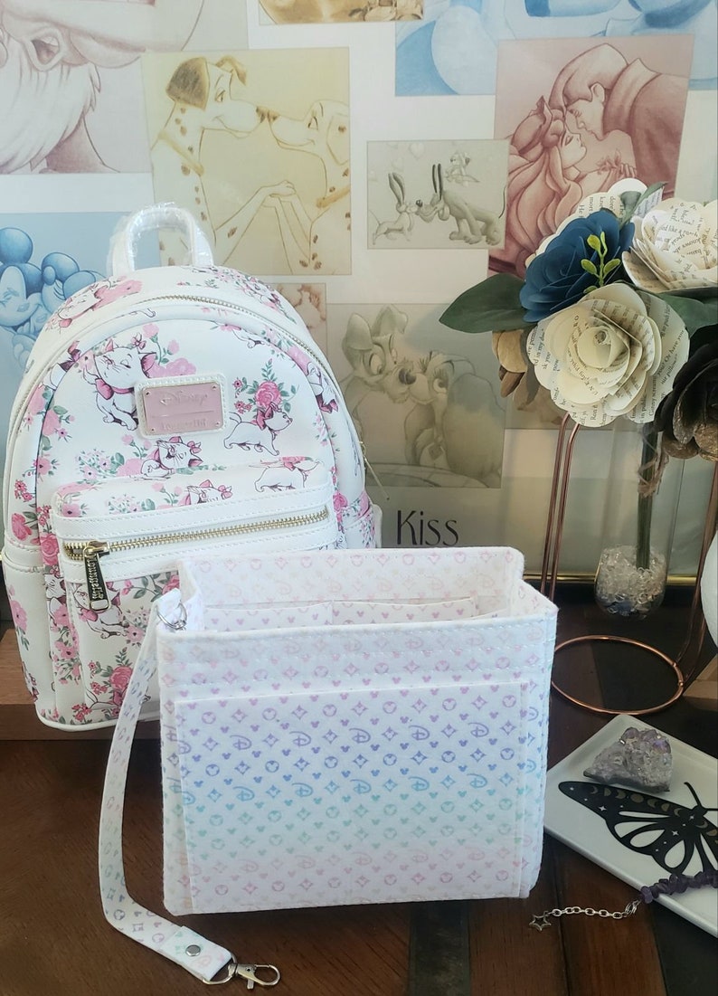 May include: White backpack with a Marie the Cat design, featuring pink flowers and a gold zipper. A matching organizer with a gradient pattern and a detachable strap sits in front. The background includes Disney art.