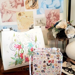 May include: White tote bag with a colorful illustration of a mermaid surrounded by flowers. A smaller organizer bag with a strap features a pattern of cartoon characters. Artwork of Disney characters is in the background.