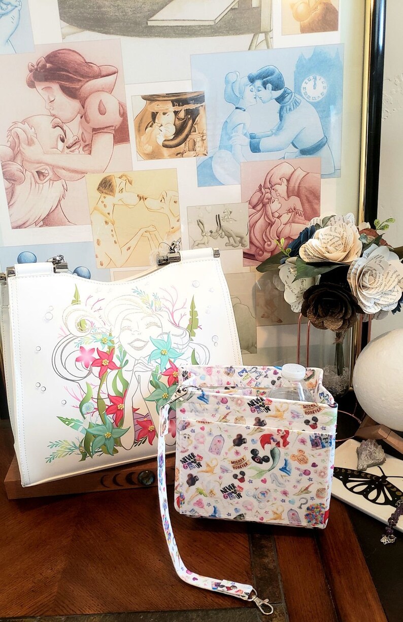 May include: White tote bag with a colorful illustration of a mermaid surrounded by flowers. A smaller organizer bag with a strap features a pattern of cartoon characters. Artwork of Disney characters is in the background.