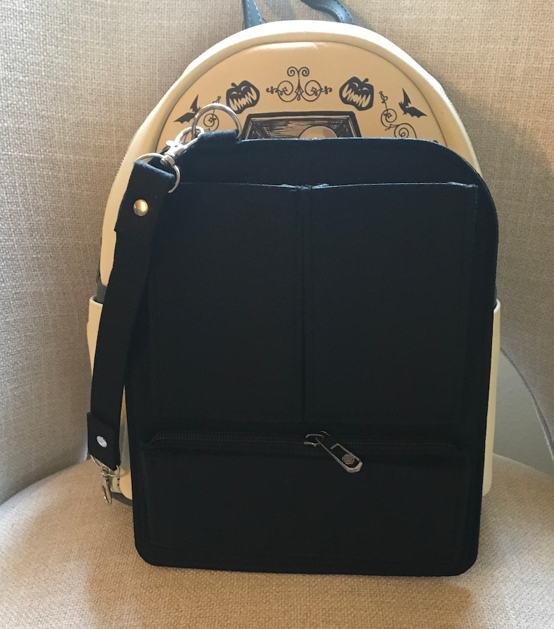 May include: Black backpack with a front zippered pocket and a detachable black strap. The backpack has a cream-colored top with a Halloween-themed design featuring pumpkins and bats.