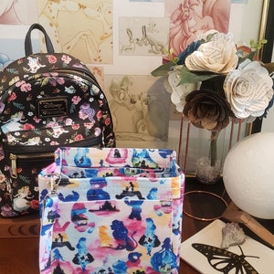 May include: A black Disney Loungefly backpack with a floral pattern and a wristlet with a colorful silhouette design. The wristlet features a detachable strap and a zipper closure. The background includes framed Disney art and paper flowers.