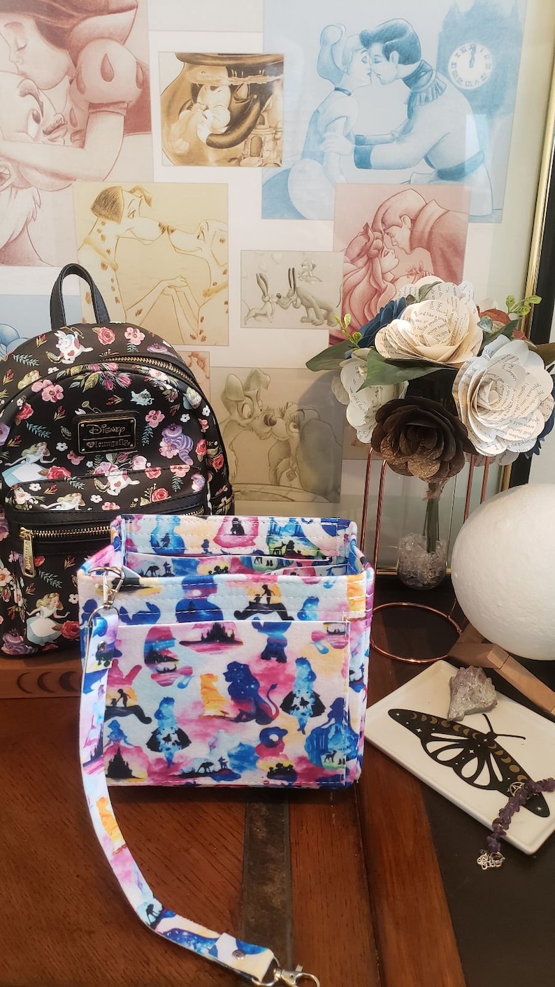May include: A black Disney Loungefly backpack with a floral pattern and a wristlet with a colorful silhouette design. The wristlet features a detachable strap and a zipper closure. The background includes framed Disney art and paper flowers.