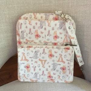 May include: A white crossbody bag with a whimsical pattern featuring Winnie the Pooh and friends. The bag has multiple pockets, a zippered compartment, and a detachable strap. The design includes images of Pooh, Eeyore, and Tigger.