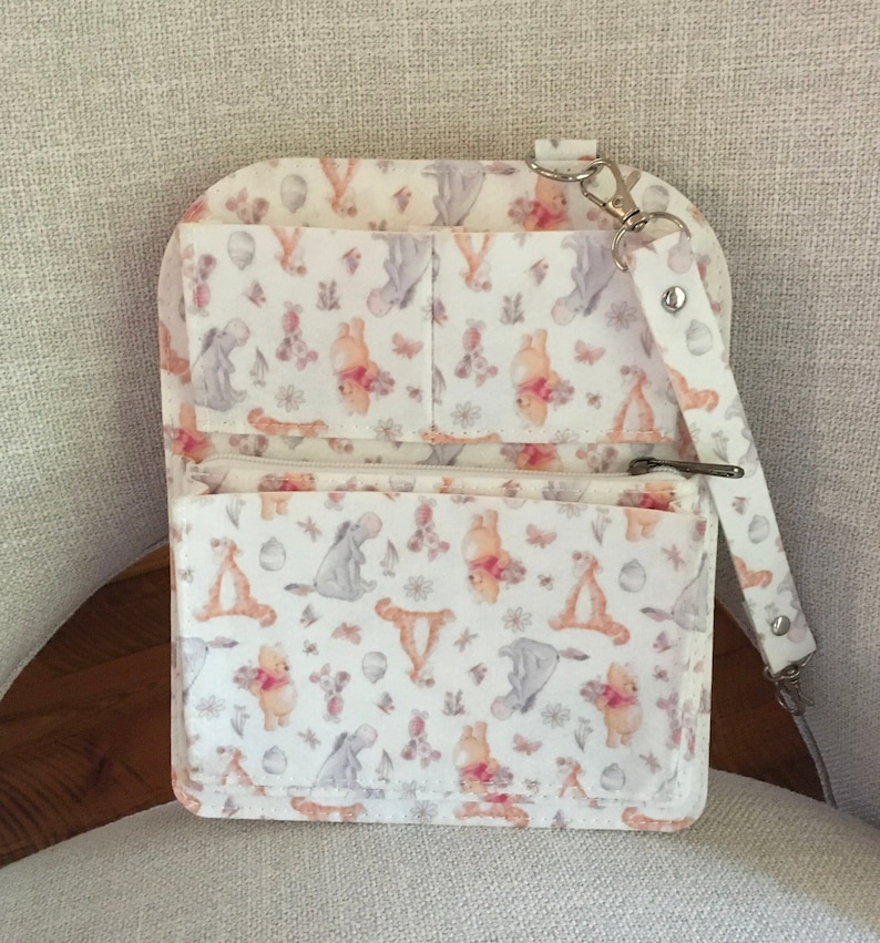 May include: A white crossbody bag with a whimsical pattern featuring Winnie the Pooh and friends. The bag has multiple pockets, a zippered compartment, and a detachable strap. The design includes images of Pooh, Eeyore, and Tigger.