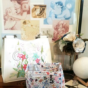 May include: White tote bag with a floral design and a patterned interior organizer. The organizer features a variety of cartoon characters. Artwork of Disney characters kissing is in the background.