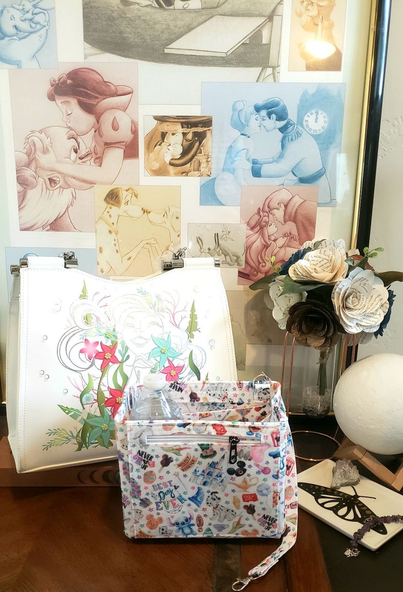 May include: White tote bag with a floral design and a patterned interior organizer. The organizer features a variety of cartoon characters. Artwork of Disney characters kissing is in the background.