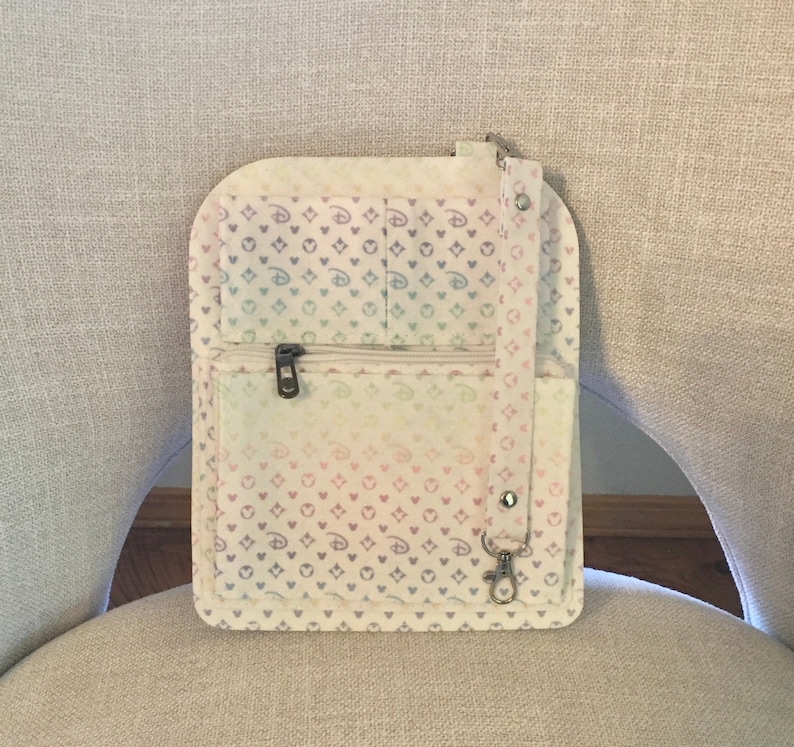 May include: A pastel-colored crossbody bag with a repeating pattern of hearts and Mickey Mouse silhouettes. The bag features a zippered pocket, a small pocket, and a silver-tone clasp. The bag is made of a soft, fabric material.