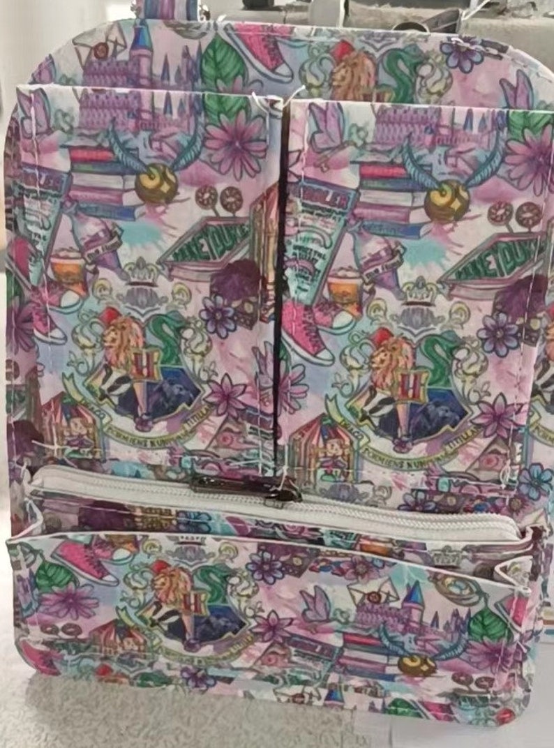 May include: A multi-pocket backpack with a colorful pattern featuring elements from the Harry Potter series. The design includes images of the Hogwarts crest, books, sneakers, and flowers. The backpack has a white zipper and multiple compartments.