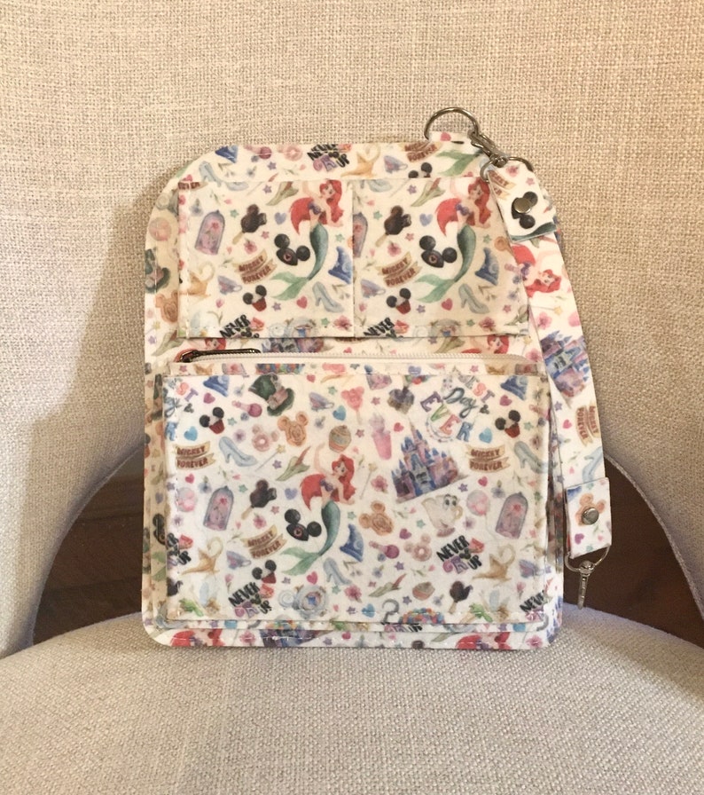 May include: A white crossbody bag with a colorful pattern featuring Disney characters and icons. The bag has a zippered front pocket, multiple compartments, and a detachable strap. The pattern includes images of Ariel, Mickey Mouse, and Cinderella's castle.