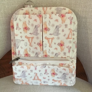 May include: A white wallet with a zipper closure and multiple pockets. The wallet features a repeating pattern of Winnie the Pooh characters, flowers, and butterflies in soft pastel colors. A silver key ring is attached to the top.