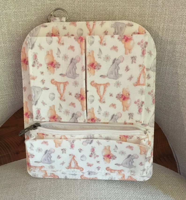 May include: A white wallet with a zipper closure and multiple pockets. The wallet features a repeating pattern of Winnie the Pooh characters, flowers, and butterflies in soft pastel colors. A silver key ring is attached to the top.